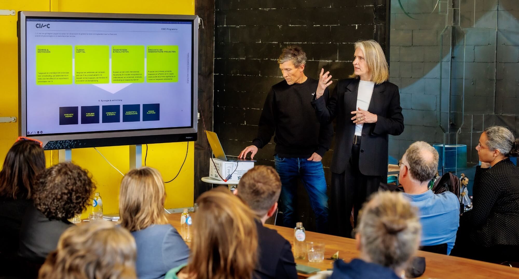 Two people are giving a presentation. On the left of the image, there is a screen displaying CIIIC. In the foreground, a group of people is seen from the back, listening to the story.